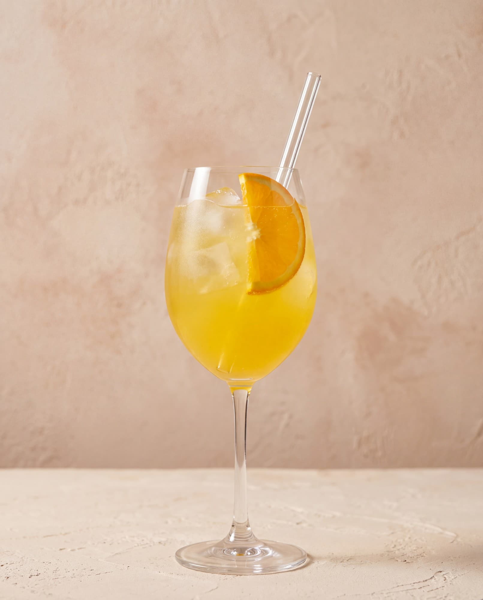 Elisira Spritz sur glace dans un grand verre à pied, avec une rondelle d'orange et une paille en verre, devant un mur de plâtre rosé